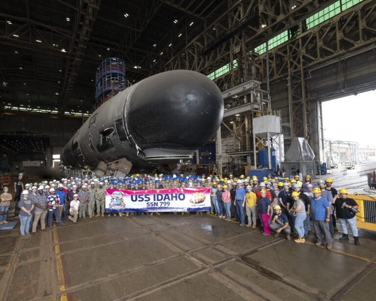 A Virginia-class submarine Idaho sits in a warehouse, behind a crowd of people holding a sigh that says “USS Idaho” at General Dynamics Electric Boat’s shipyard in Groton, Connecticut.