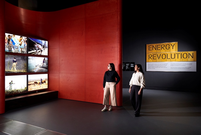 Two visitors in a red gallery look at a wall of six screens.