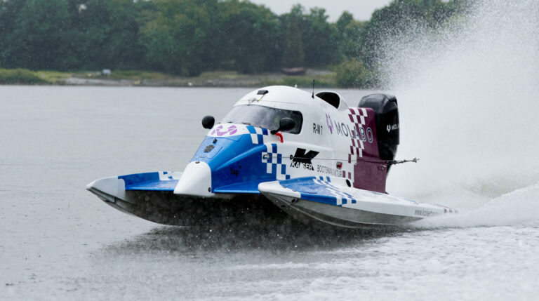 Electric Formula 4 boat racing across an inland waterway at high speed.