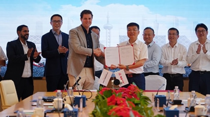 Two men shake hands, while holding a signed contract, at a signing ceremony full of clapping men.
