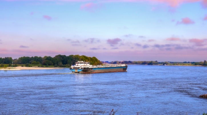 A small vessel sails along a river at dusk, with the shore visible in the background, with trees and a beach.
