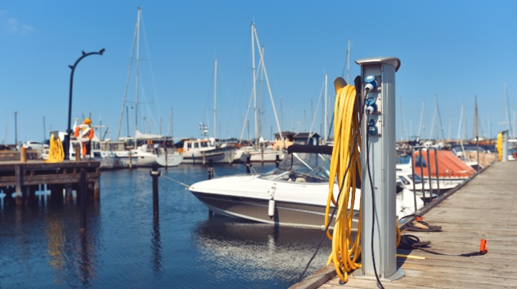 An electric pole sockets stands on a marina pier, with boats in the background.
