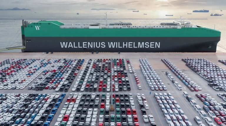 A large Wallenius Wilhelmsen container vessel moored at a container port. The sun is low in the sky in the background.