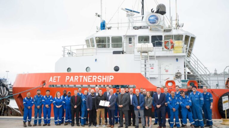 AET and Fleetzero representatives stand in front of the plug-in hybrid-electric lightering vessel that will be retrofitted as part of a new agreement. the vessel is orange and white and the staff wear suits or blue workwear.