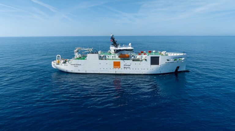 A white Orange Marine cable repair vessel with orange company branding. The vessel is sailing in calm blue waters.