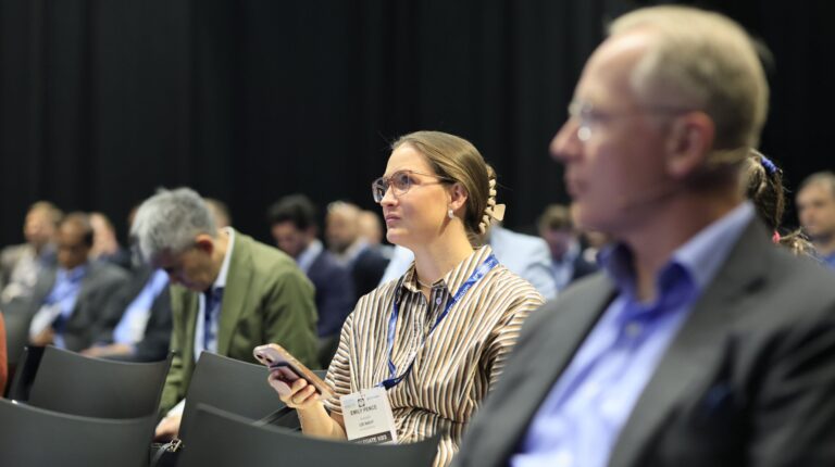 Conference attendees sit in rows listening to a speaker. The wall in the background is black.