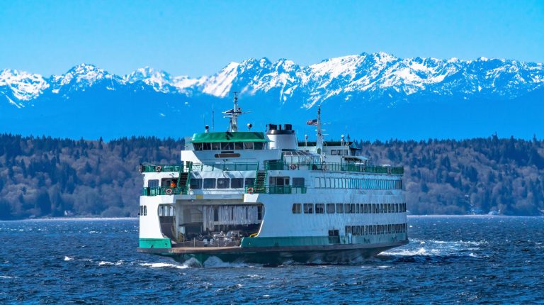 A Washington State Ferry sails through the water with mountains visible in the background.